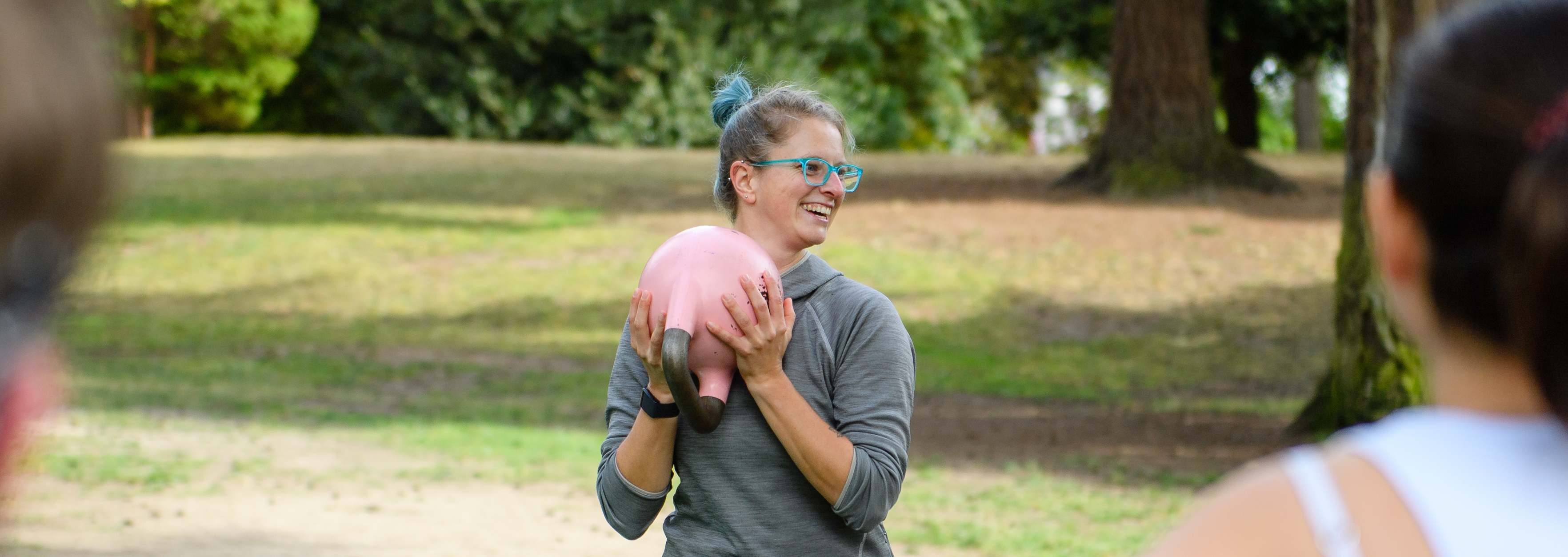 Bert, a white person with colorful hair, stands outside holding a kettlebell and smiling in front of a group of people.